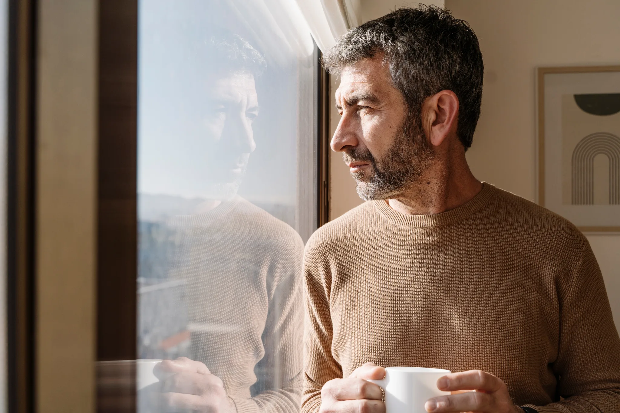 Man holding a coffee mug, looking out a sunlit window.